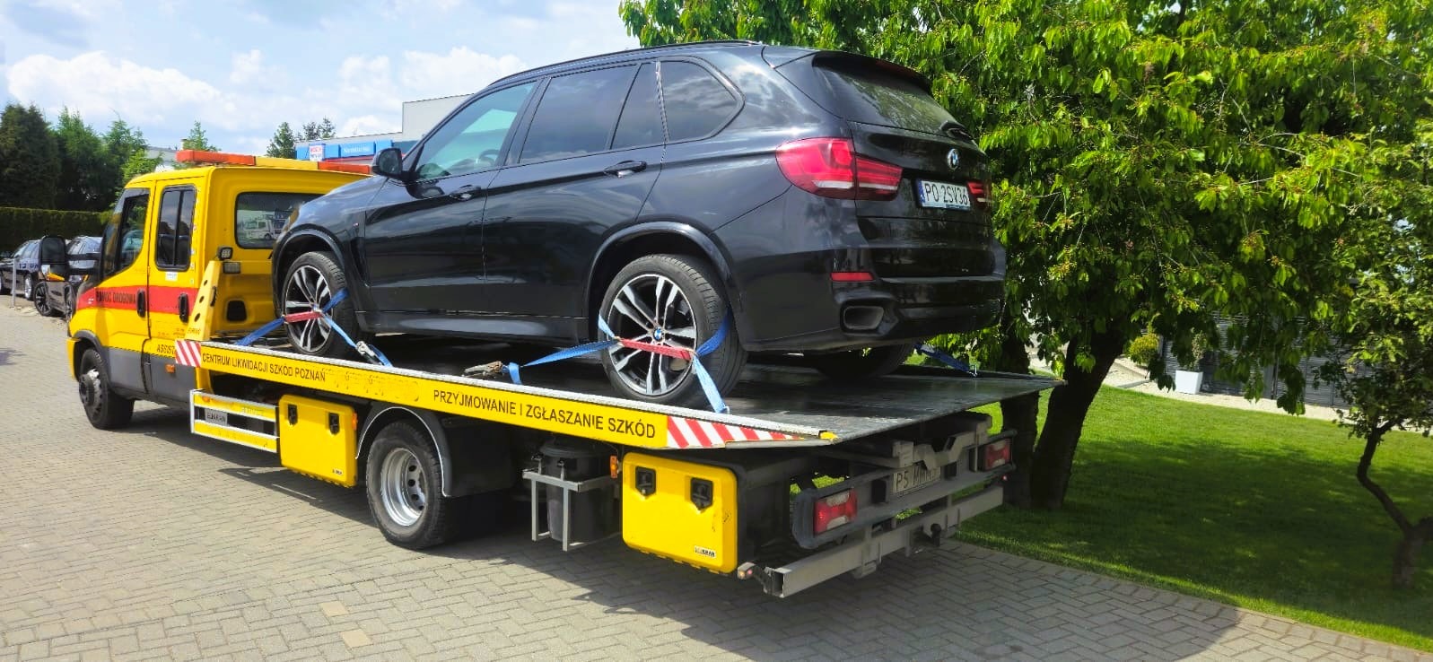 Czarny SUV BMW na żółtej lawecie pomocy drogowej, zabezpieczony pasami, z widocznym numerem rejestracyjnym na tle zieleni.