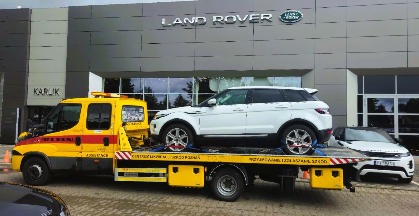 Żółta laweta z białym Land Roverem na tle salonu Karlik. Na aucie napis: Pomoc Drogowa. Drugi Range Rover stoi obok. Centrum Likwidacji Szkód Poznań.