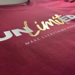 MAKT WIKTOR JAKUBOWSKI - Burgundowa koszulka z napisem 'Unlimited' w kolorze złotym i białym, leżąca płasko, ukazująca detale nadruku i fakturę materiału.