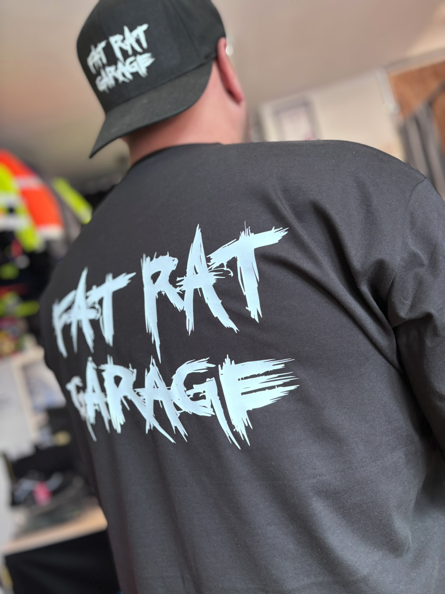 Czarna koszulka i czapka z białym nadrukiem 'FAT RAT GARAGE', widok z tyłu na osobie. Grafika w stylu graffiti. Elementy garderoby z nadrukiem.