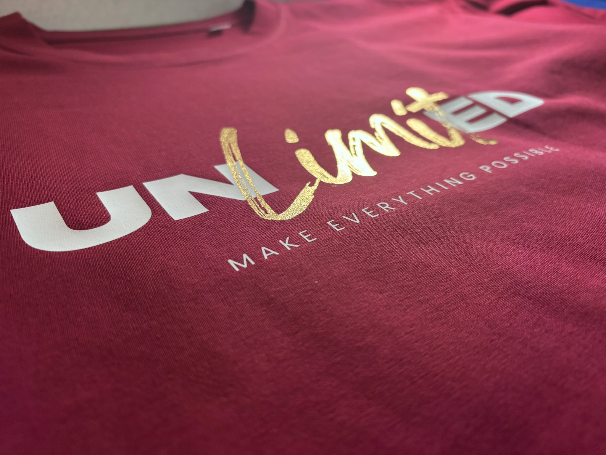 Burgundowa koszulka z napisem 'Unlimited' w kolorze złotym i białym, leżąca płasko, ukazująca detale nadruku i fakturę materiału.