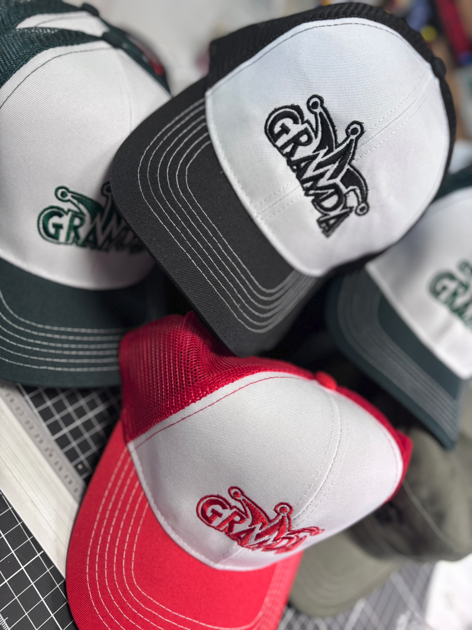 Stos czapek z daszkiem: czerwona, czarna i zielona, każda z haftowanym logo 'Granda' w różnych kolorach. Widoczne detale siateczki i szwy czapek.