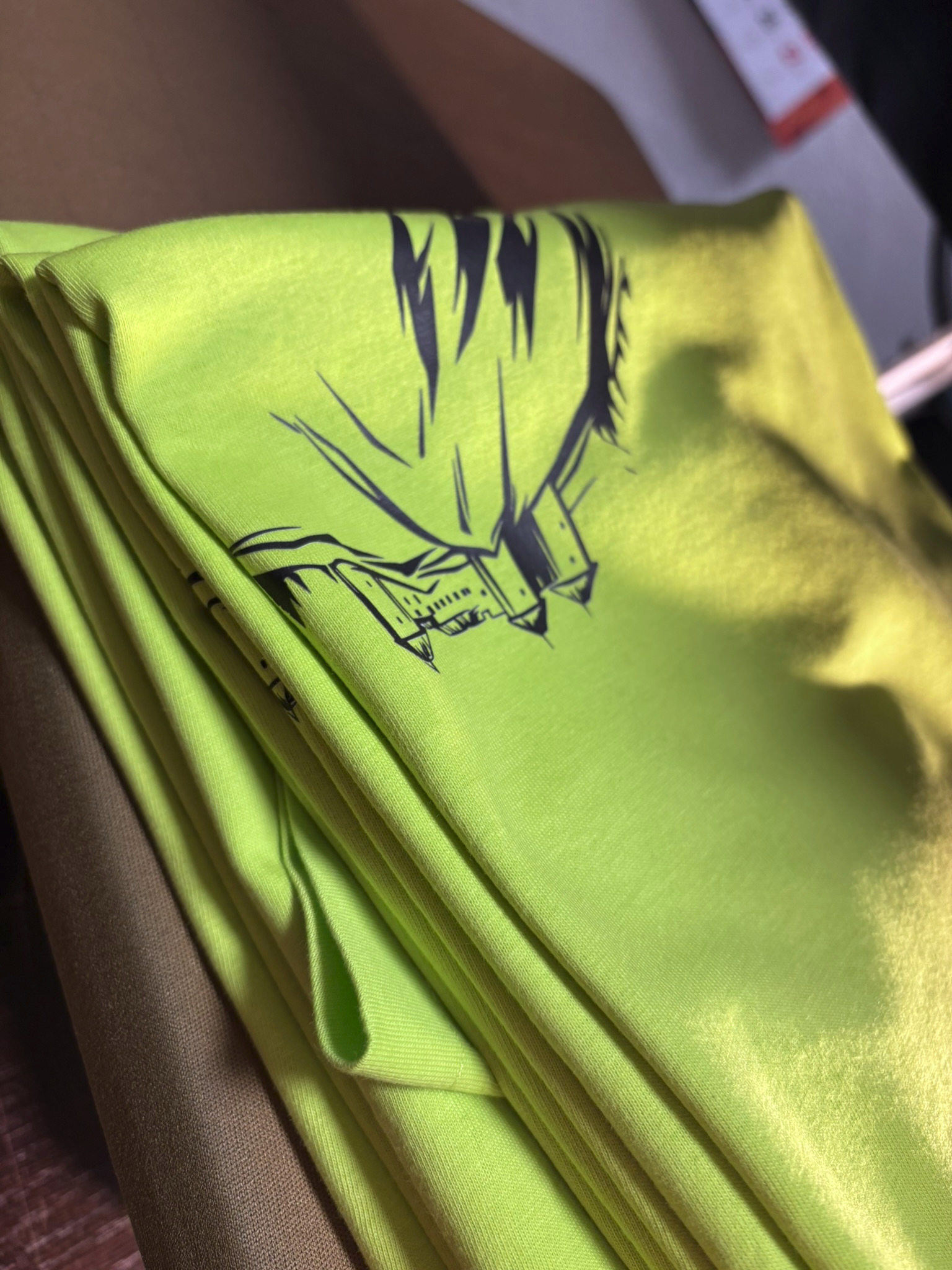 Stos neonowo zielonych koszulek z czarnym nadrukiem graficznym, ułożonych na sobie. W tle widoczny fragment brązowej koszulki i rozmazane tło.