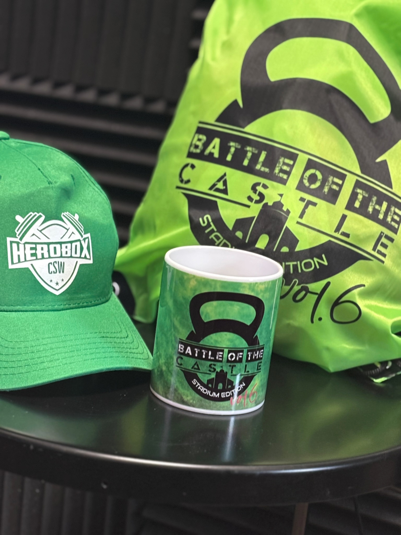 Zielona czapka z logo HeroBox CSW, kubek i torba z nadrukiem Battle of the Castle, prezentujące gadżety firmowe na ciemnym stole.