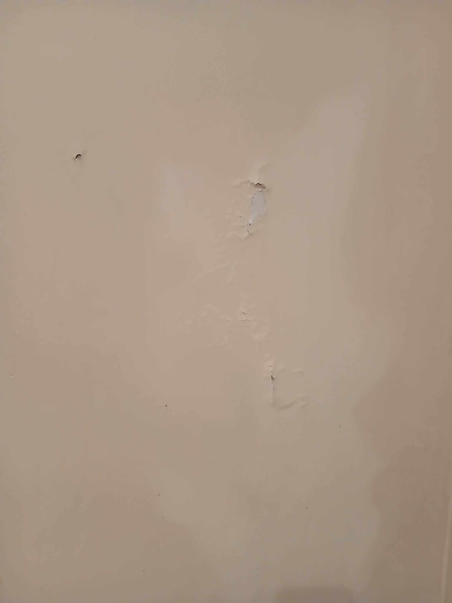 Ściana w beżowym odcieniu z widocznymi uszkodzeniami i odpryskami farby, ukazująca stan przed malowaniem. Nierówna powierzchnia, widoczne ślady po naprawach.