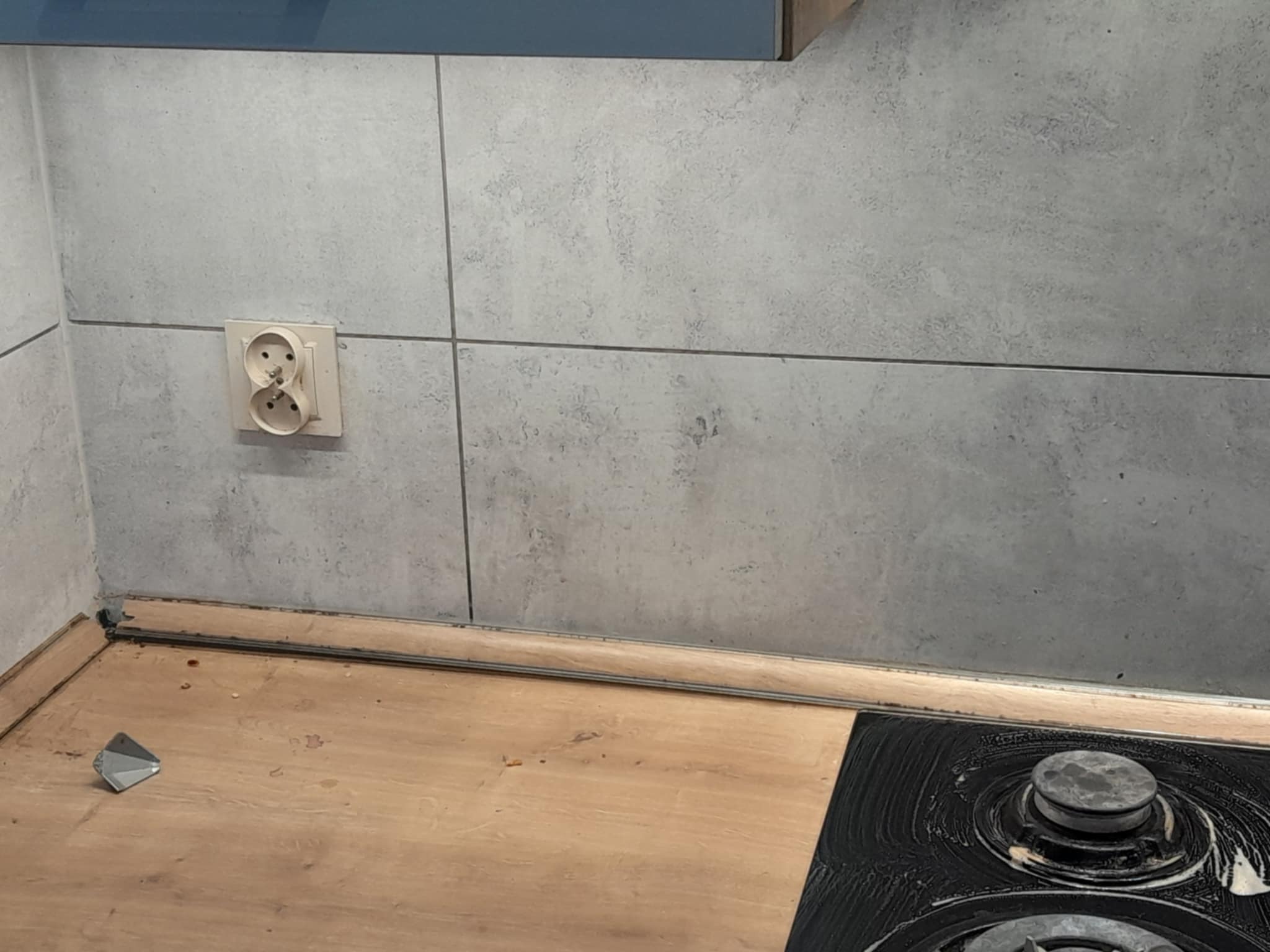 Brudna płyta kuchenna z resztkami jedzenia, widoczna szpara między blatem a ścianą, gniazdko elektryczne. Stan przed sprzątaniem.