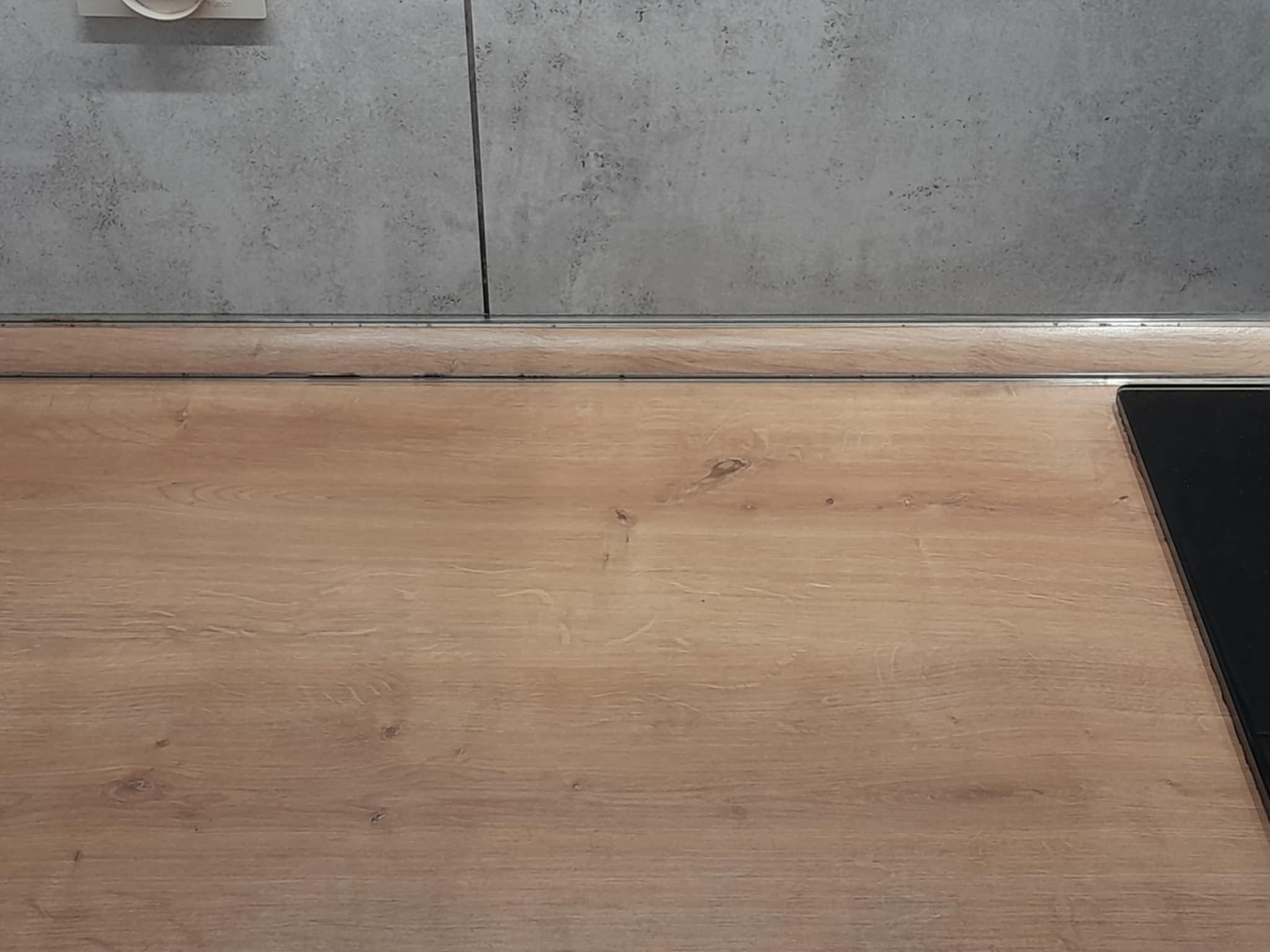 Wykończona, czysta drewniana powierzchnia blatu kuchennego z szarą ścianą w tle, po sprzątaniu. Fragment płyty indukcyjnej w prawym dolnym rogu.