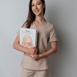 Kosmetologia Agnieszka Gomółka-Szul - Uśmiechnięta brunetka w beżowym uniformie trzyma atlas anatomii twarzy, prezentując wiedzę z zakresu medycyny estetycznej i zabiegów kosmetycznych.