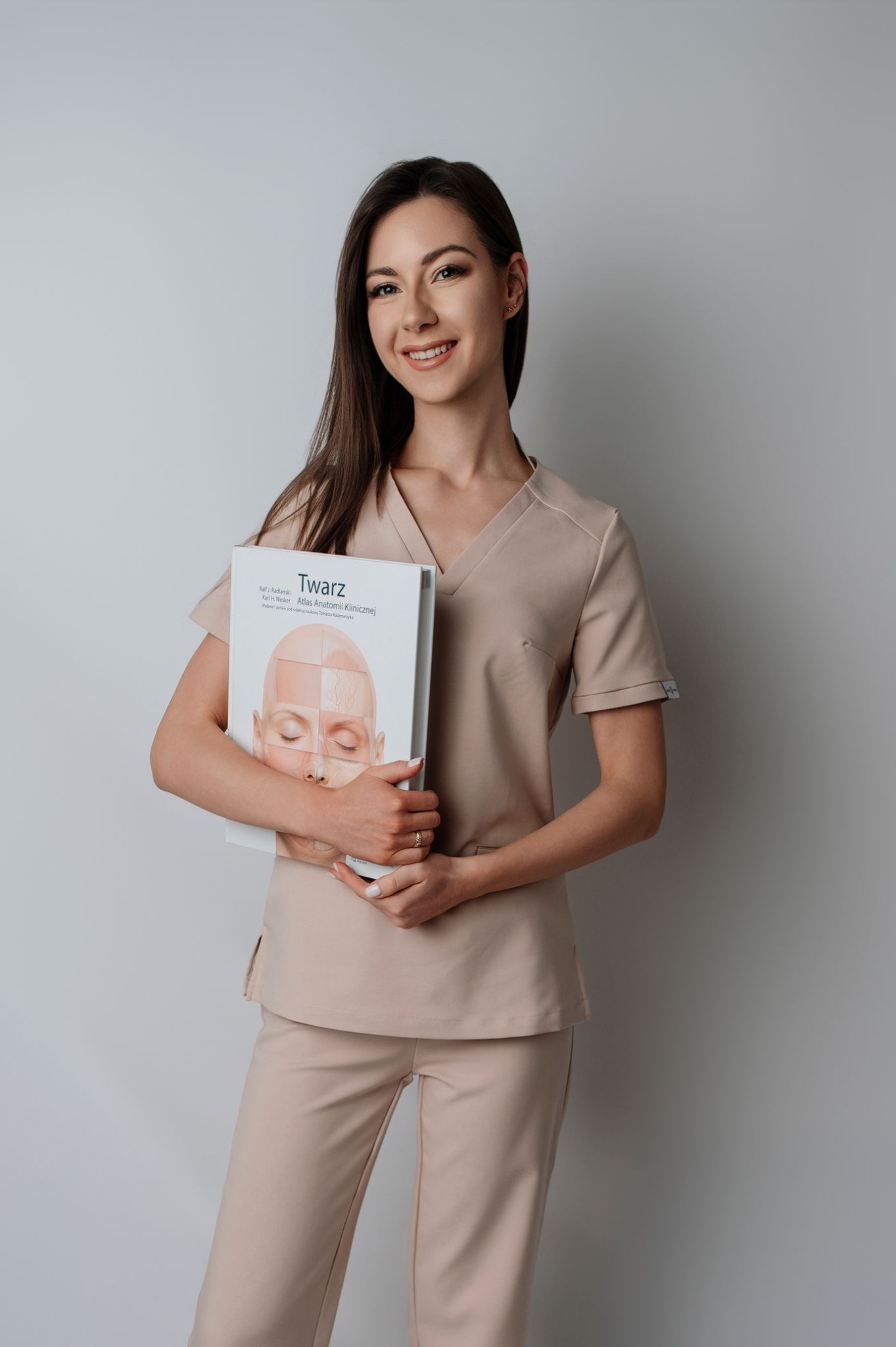 Uśmiechnięta brunetka w beżowym uniformie trzyma atlas anatomii twarzy, prezentując wiedzę z zakresu medycyny estetycznej i zabiegów kosmetycznych.