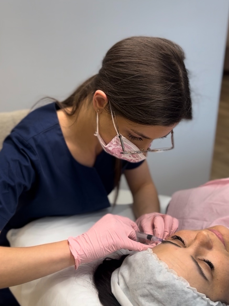 Kosmetolog w różowych rękawiczkach wykonuje zabieg na brwiach pacjentki, używając strzykawki. Kobieta w okularach i maseczce ochronnej skupiona na precyzyjnej aplikacji.
