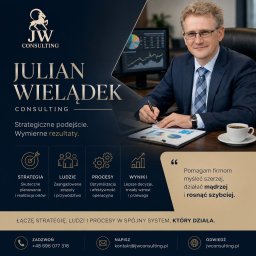 Julian Wielądek - consulting - Uśmiechnięty konsultant w okularach i garniturze, piszący na dokumencie z wykresami przy biurku z laptopem i monitorem, w tle logo firmy JW Consulting.