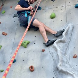 mateuszmis.com - Uśmiechnięty mężczyzna z aparatem w uprzęży wspinaczkowej na ściance, ujęcie z dołu, lina i kolorowe chwyty wspinaczkowe.