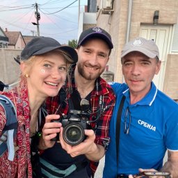 mateuszmis.com - Ekipa filmowa w plenerze: uśmiechnięta blondynka z brokatem, mężczyzna z kamerą Lumix GHS5 i mężczyzna w koszulce Serbia. W tle zabudowa mieszkalna.