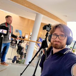 mateuszmis.com - Ekipa filmowa podczas nagrywania wywiadu na tle ceglanej ściany, z widoczną kamerą na statywie i oświetleniem. W tle uczestnicy konferencji.