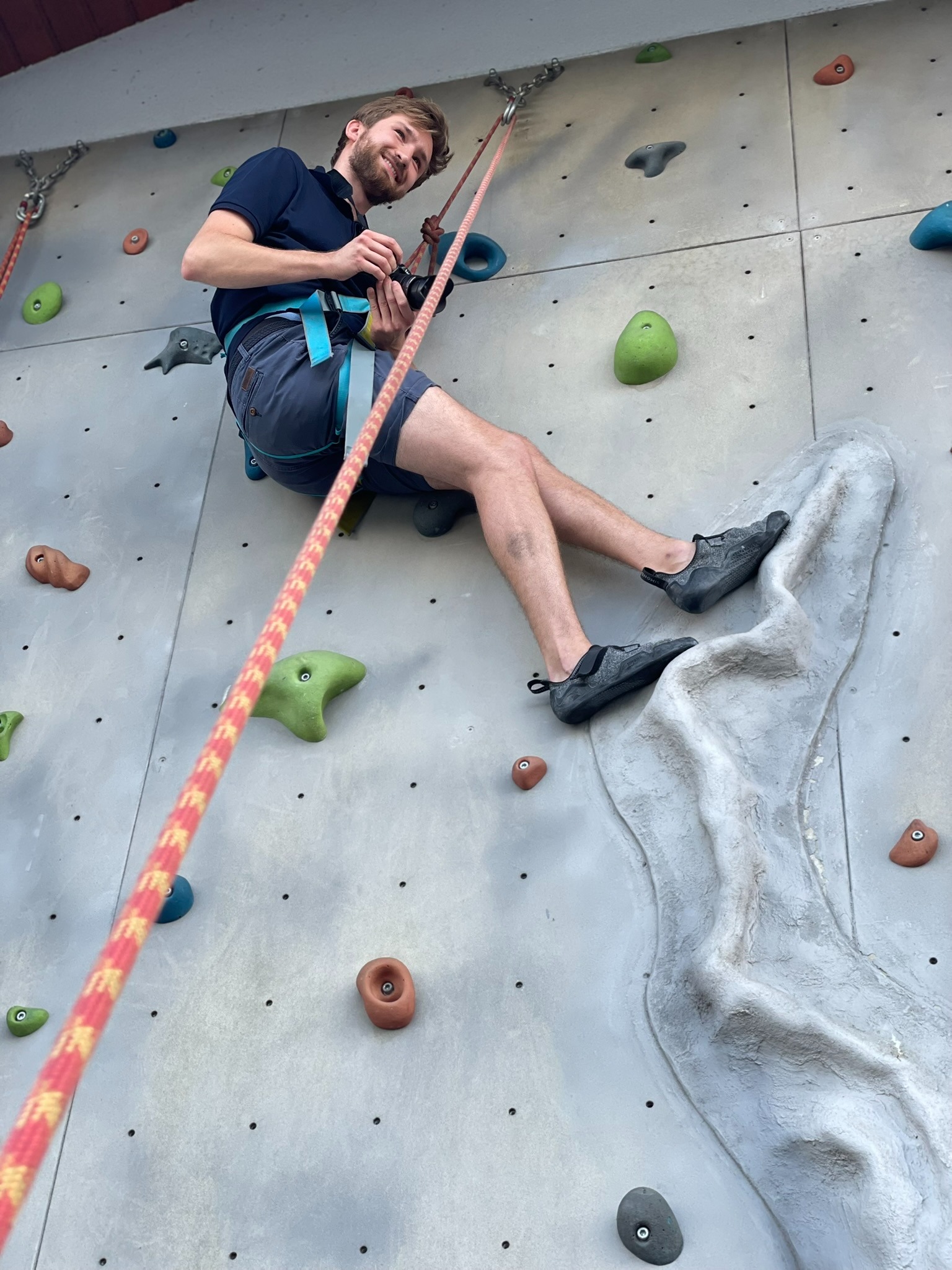 Uśmiechnięty mężczyzna z aparatem w uprzęży wspinaczkowej na ściance, ujęcie z dołu, lina i kolorowe chwyty wspinaczkowe.