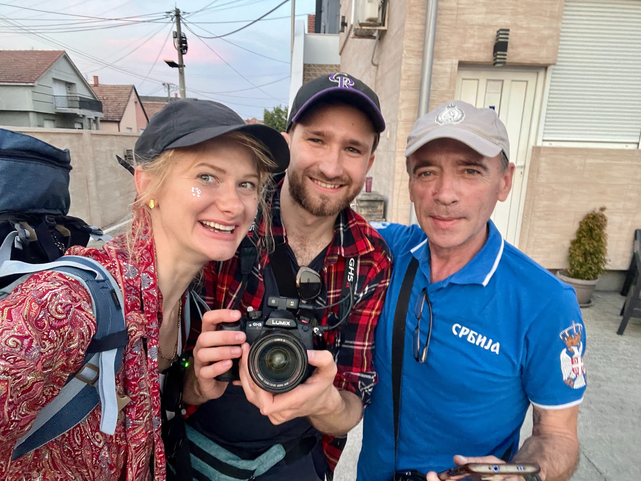 Ekipa filmowa w plenerze: uśmiechnięta blondynka z brokatem, mężczyzna z kamerą Lumix GHS5 i mężczyzna w koszulce Serbia. W tle zabudowa mieszkalna.