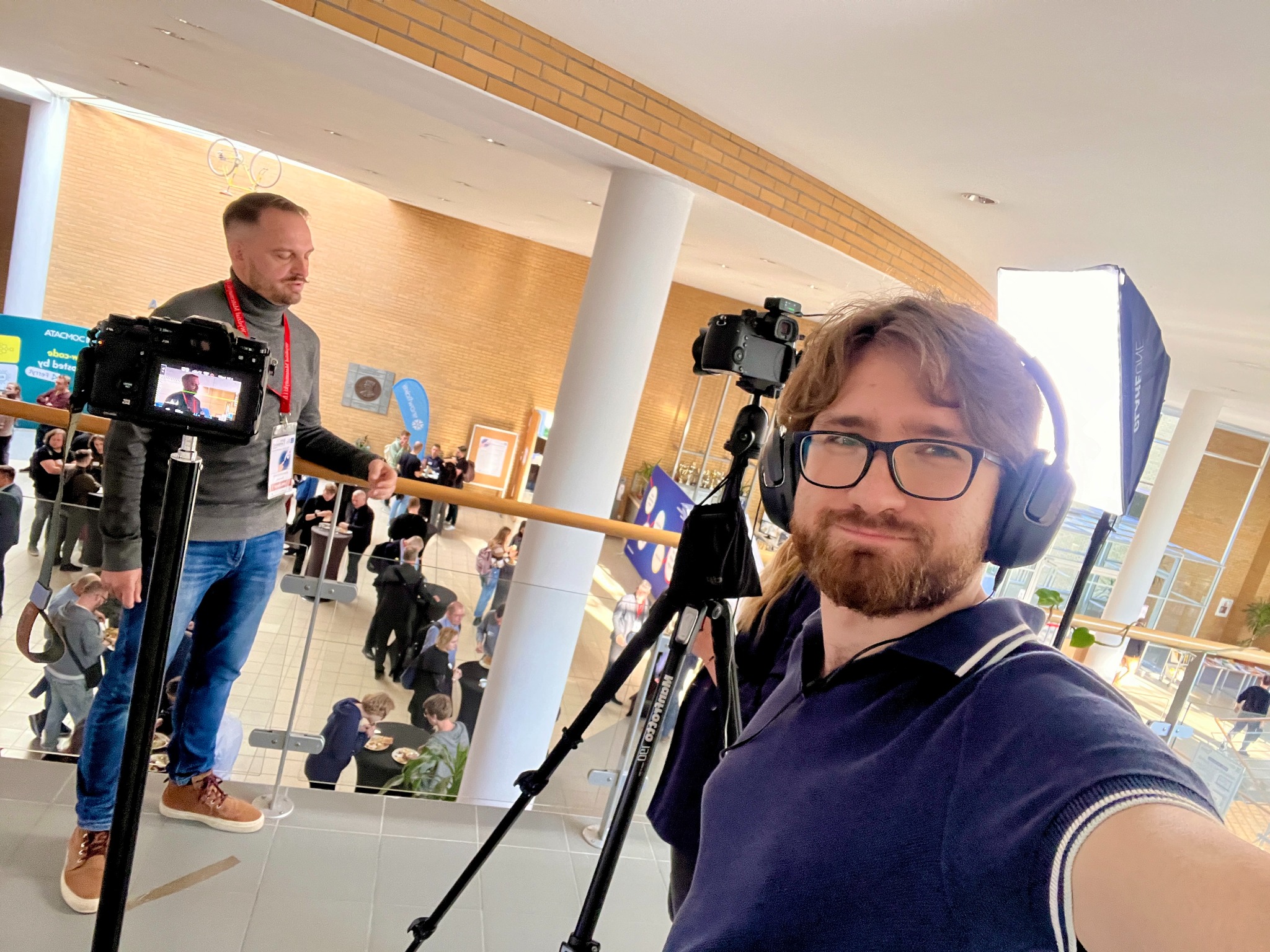 Ekipa filmowa podczas nagrywania wywiadu na tle ceglanej ściany, z widoczną kamerą na statywie i oświetleniem. W tle uczestnicy konferencji.