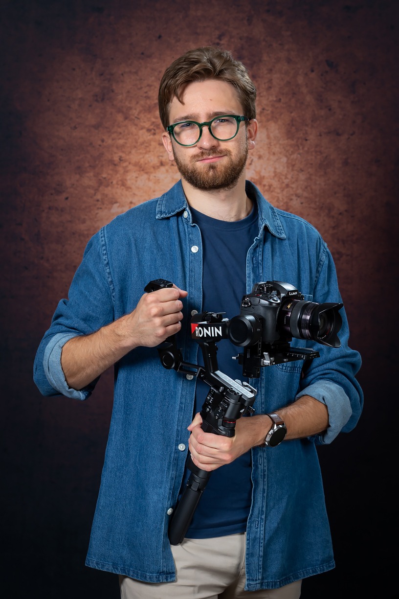 Uśmiechnięty mężczyzna w okularach prezentuje kamerę Lumix z gimbalem Ronin na brązowym tle, demonstrując sprzęt do filmowania.