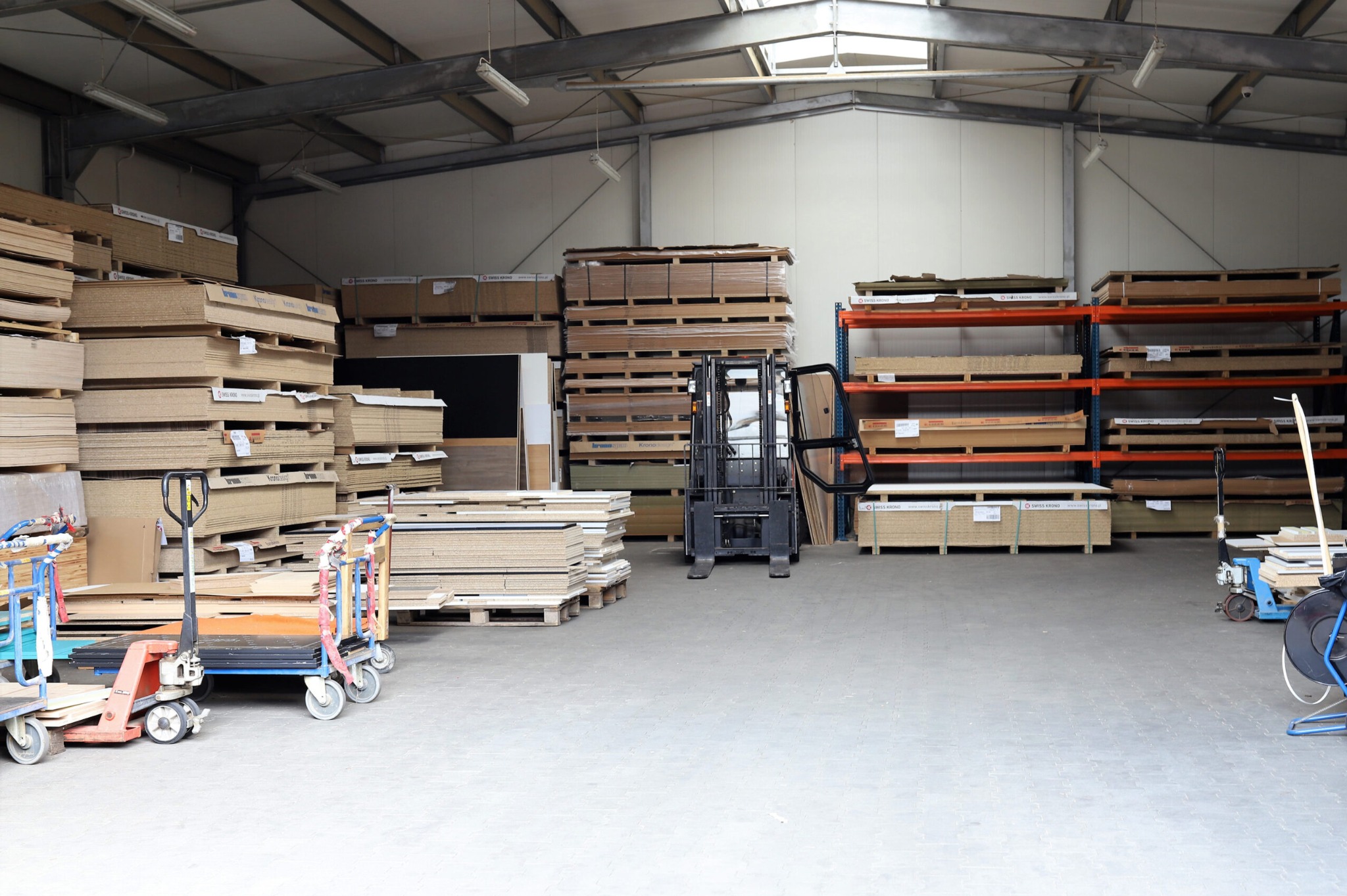 Magazyn płyt drewnianych i sklejek z wózkiem widłowym, paletami i regałami. Widoczne różne grubości i rodzaje materiałów, oświetlenie magazynowe.