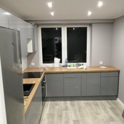 Remont - Szara kuchnia z drewnianym blatem pod oknem. Widoczna lodówka i płyta indukcyjna. Na podłodze panele. Na blacie akcesoria i butelka wody.