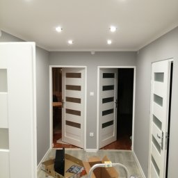 Remont - Szare wnętrze po remoncie z trzema białymi drzwiami z przeszkleniami. Na podłodze kartony, drabina. Sufit z oświetleniem punktowym.