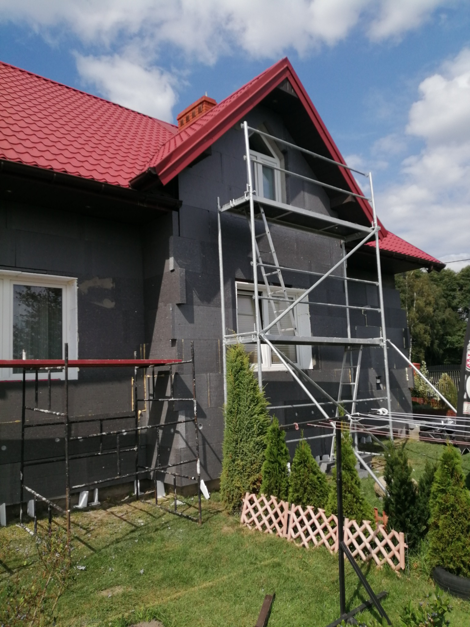 Dom jednorodzinny w trakcie ocieplania, widoczne rusztowania i styropian na elewacji. Czerwony dach kontrastuje z szarym ociepleniem. Słoneczny dzień.