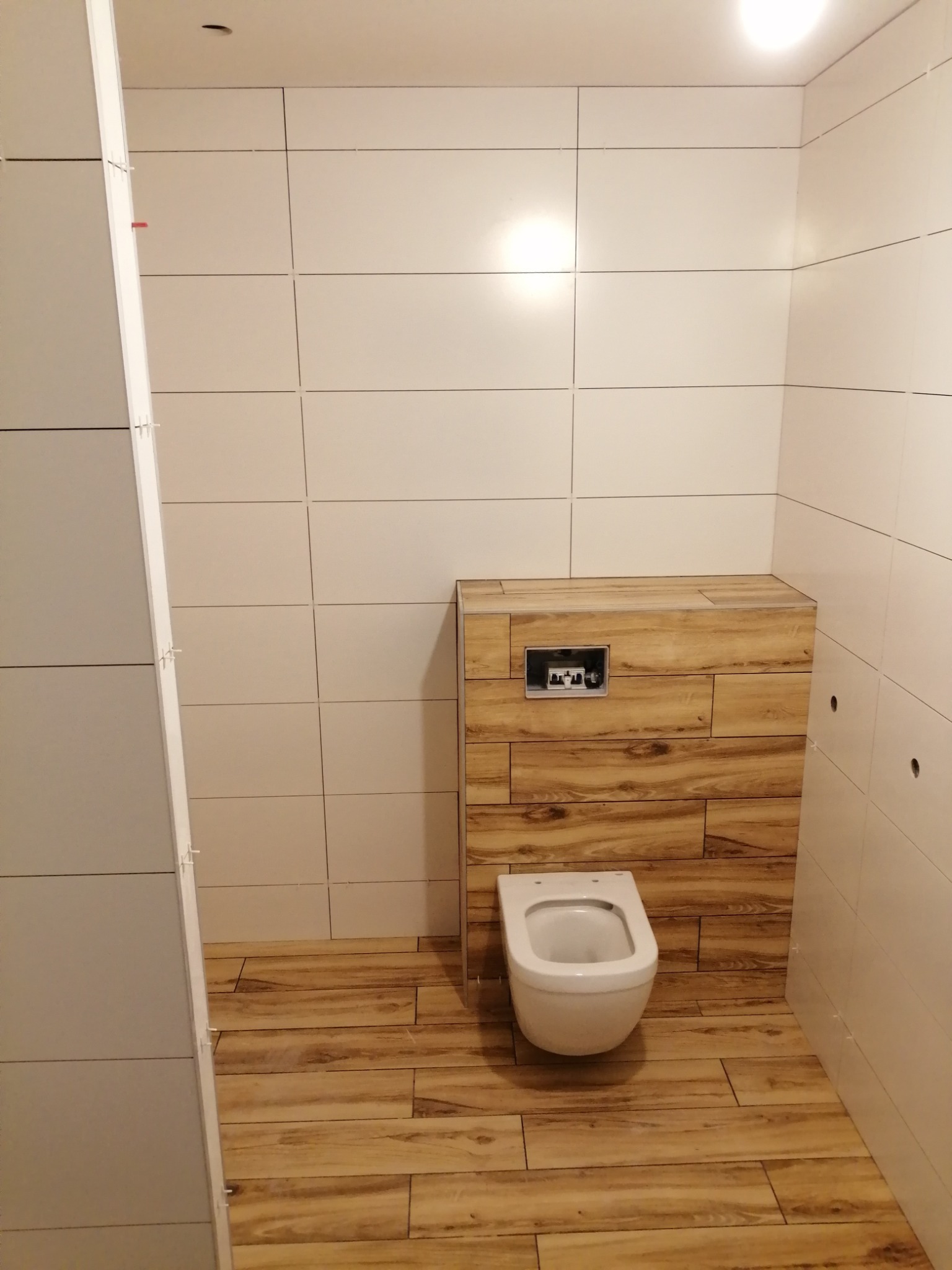 Nowoczesna łazienka z wiszącą toaletą i drewnopodobnymi płytkami na podłodze i zabudowie stelaża, kremowe płytki ścienne, minimalistyczny design.