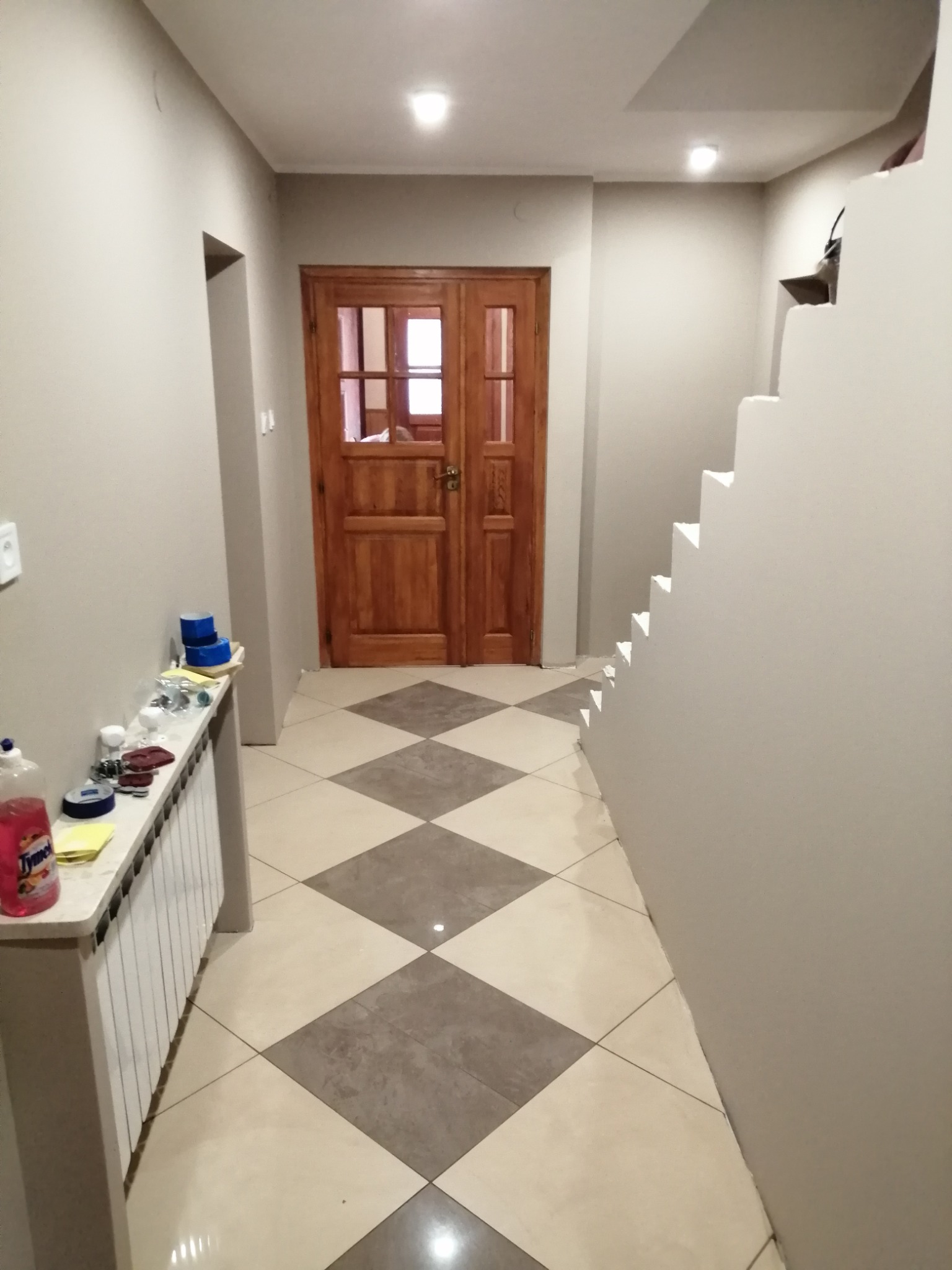 Korytarz z drewnianymi drzwiami i schodami. Podłoga wyłożona płytkami w szachownicę. Na ścianie grzejnik z akcesoriami malarskimi. Neutralna kolorystyka ścian.