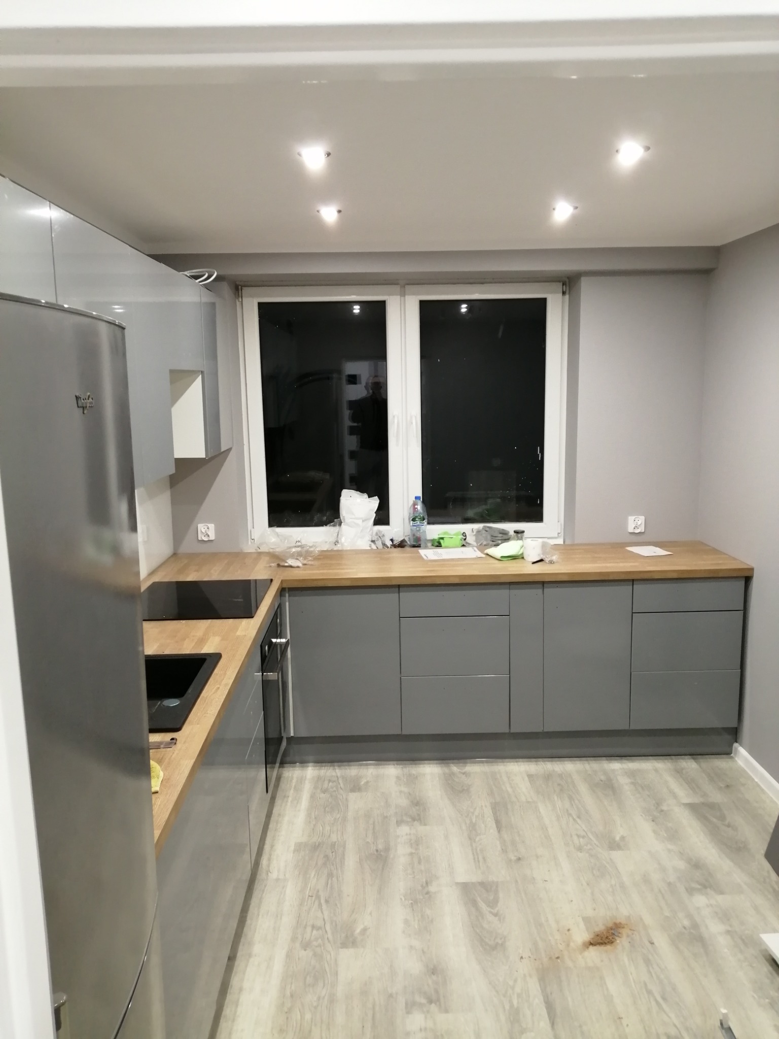 Szara kuchnia z drewnianym blatem pod oknem. Widoczna lodówka i płyta indukcyjna. Na podłodze panele. Na blacie akcesoria i butelka wody.