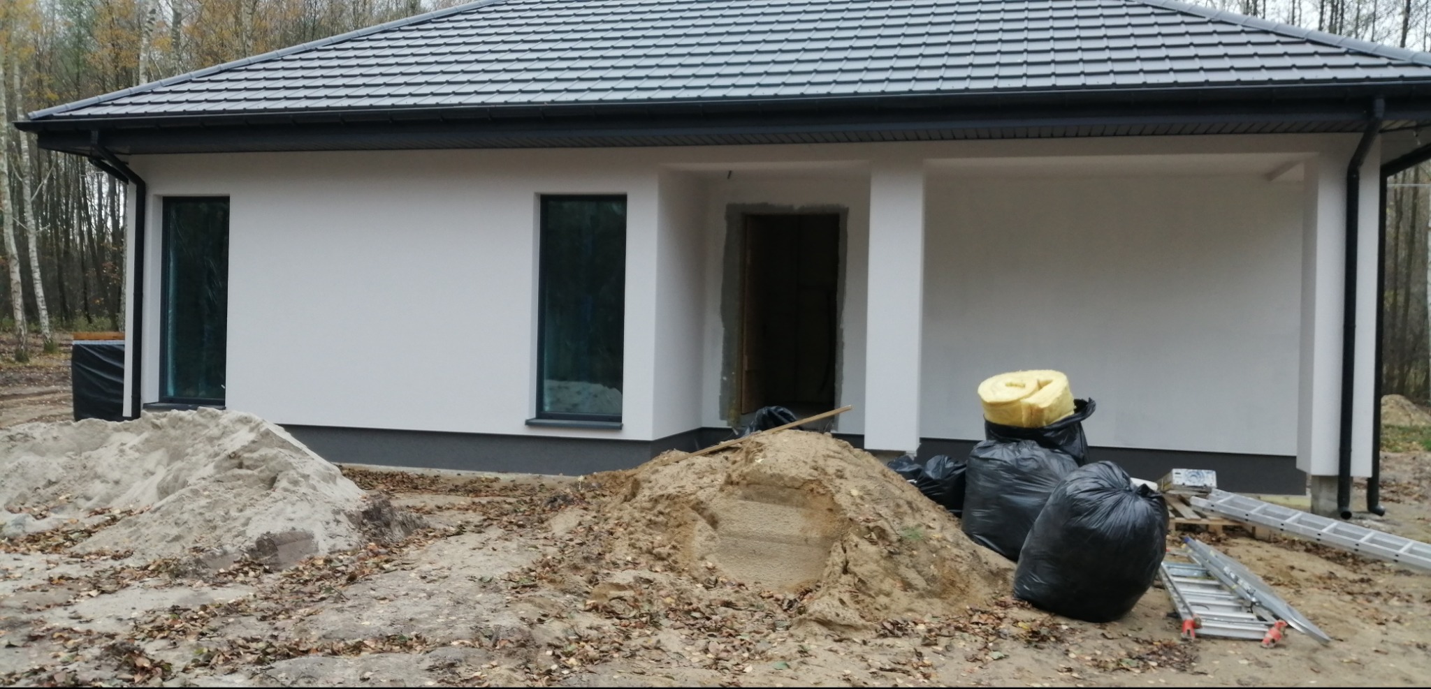 Parterowy dom z białymi ścianami i szarą dachówką w trakcie wykończenia. Na pierwszym planie piasek, worki na śmieci i drabiny.
