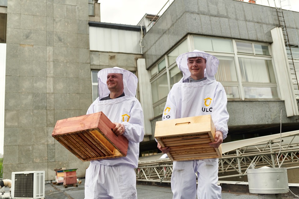Dwóch pszczelarzy w kombinezonach ULC trzyma ramki z miodem na dachu budynku. W tle widoczne ule i klimatyzator. Ujęcie z dołu, pochmurne niebo.