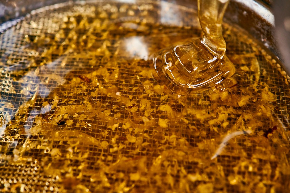 Klarowny, złocisty nektar spływa na siatkę z resztkami wosku. Makro, ciepłe światło podkreśla detale. Proces filtracji.