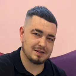 Portret mężczyzny z krótką fryzurą typu fade, w czarnej koszuli polo, uśmiechającego się do kamery na tle różowo-fioletowej ściany. Widoczny lekki zarost i srebrny łańcuszek.