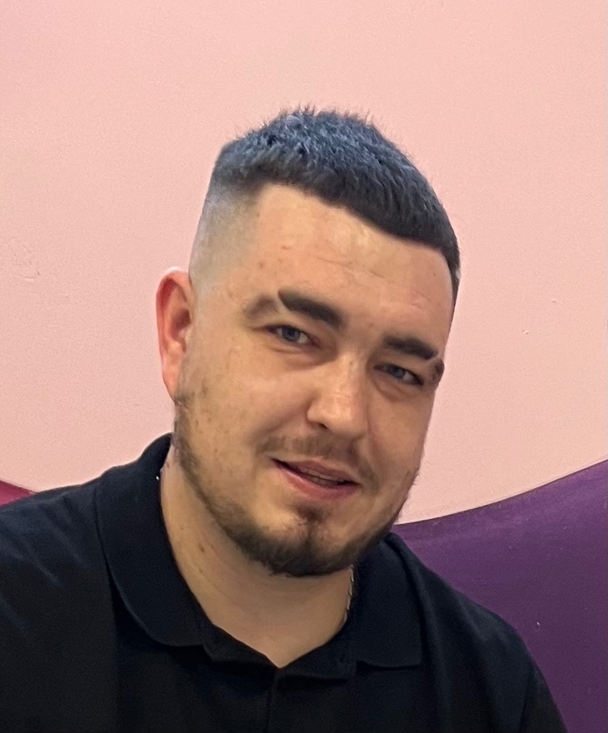 Portret mężczyzny z krótką fryzurą typu fade, w czarnej koszuli polo, uśmiechającego się do kamery na tle różowo-fioletowej ściany. Widoczny lekki zarost i srebrny łańcuszek.