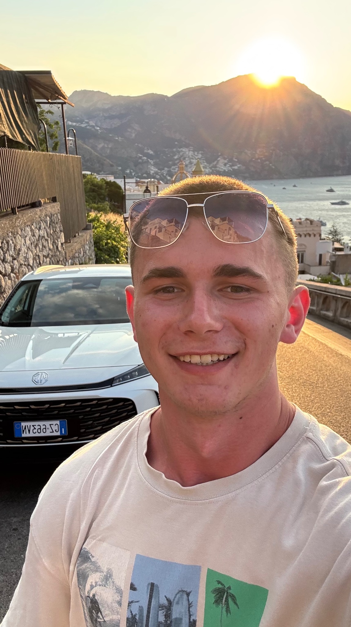 Uśmiechnięty mężczyzna w okularach przeciwsłonecznych na tle białego auta, ulicy i malowniczego krajobrazu z górami i morzem o zachodzie słońca.