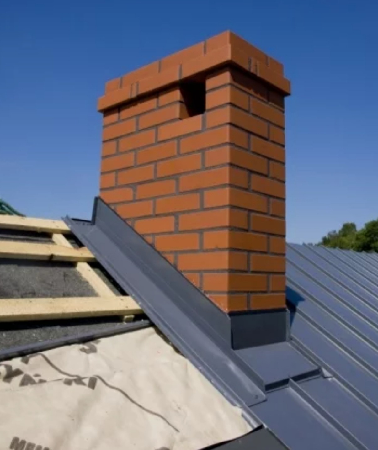 Detal dachu z ceglanym kominem i nową, szarą obróbką blacharską przy słonecznej pogodzie. Widoczne elementy konstrukcji dachu i izolacja.