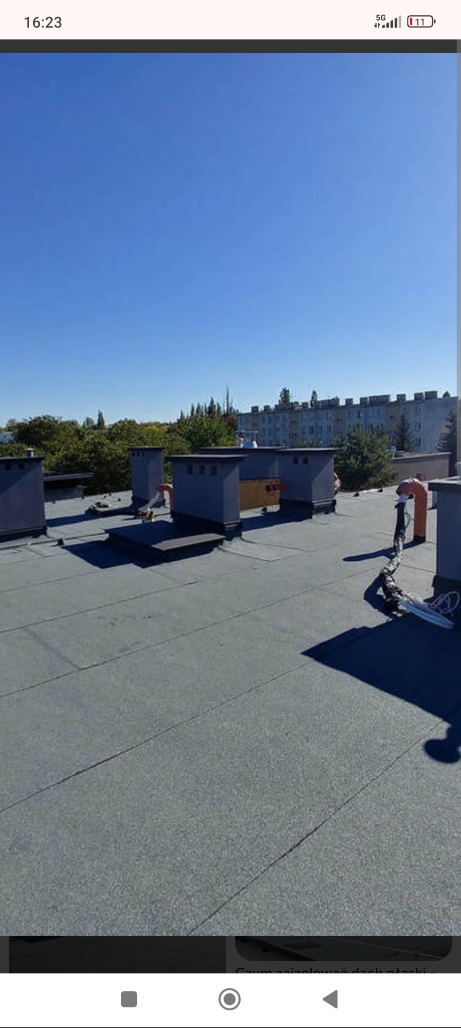 Płaski dach pokryty szarą papą termozgrzewalną z kominami wentylacyjnymi i przewodami. W tle drzewa i budynek wielorodzinny w słoneczny dzień.