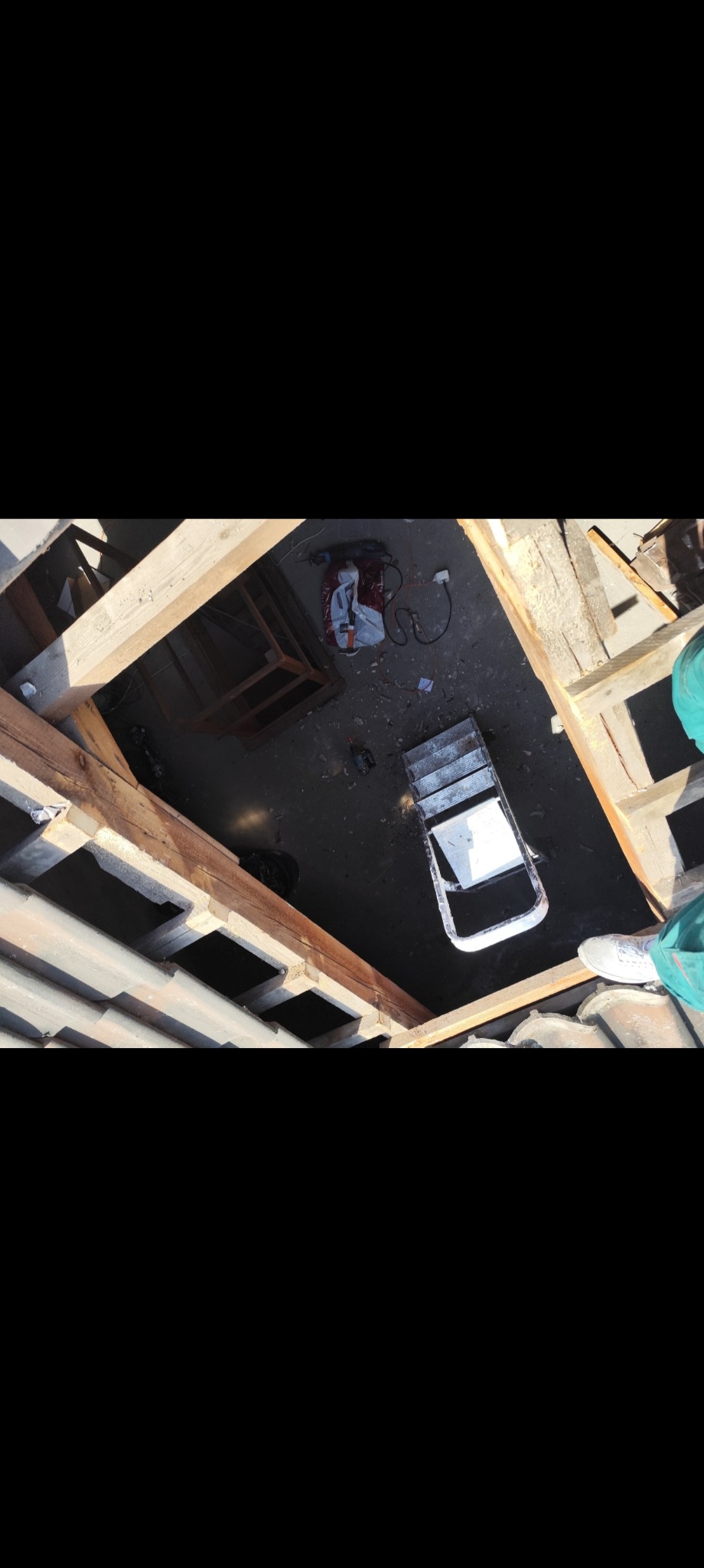 Widok z góry na rozłożony dach z odsłoniętą konstrukcją więźby, narzędzia pracy i drabinę. Fragment nogi w białym bucie widoczny w prawym dolnym rogu.