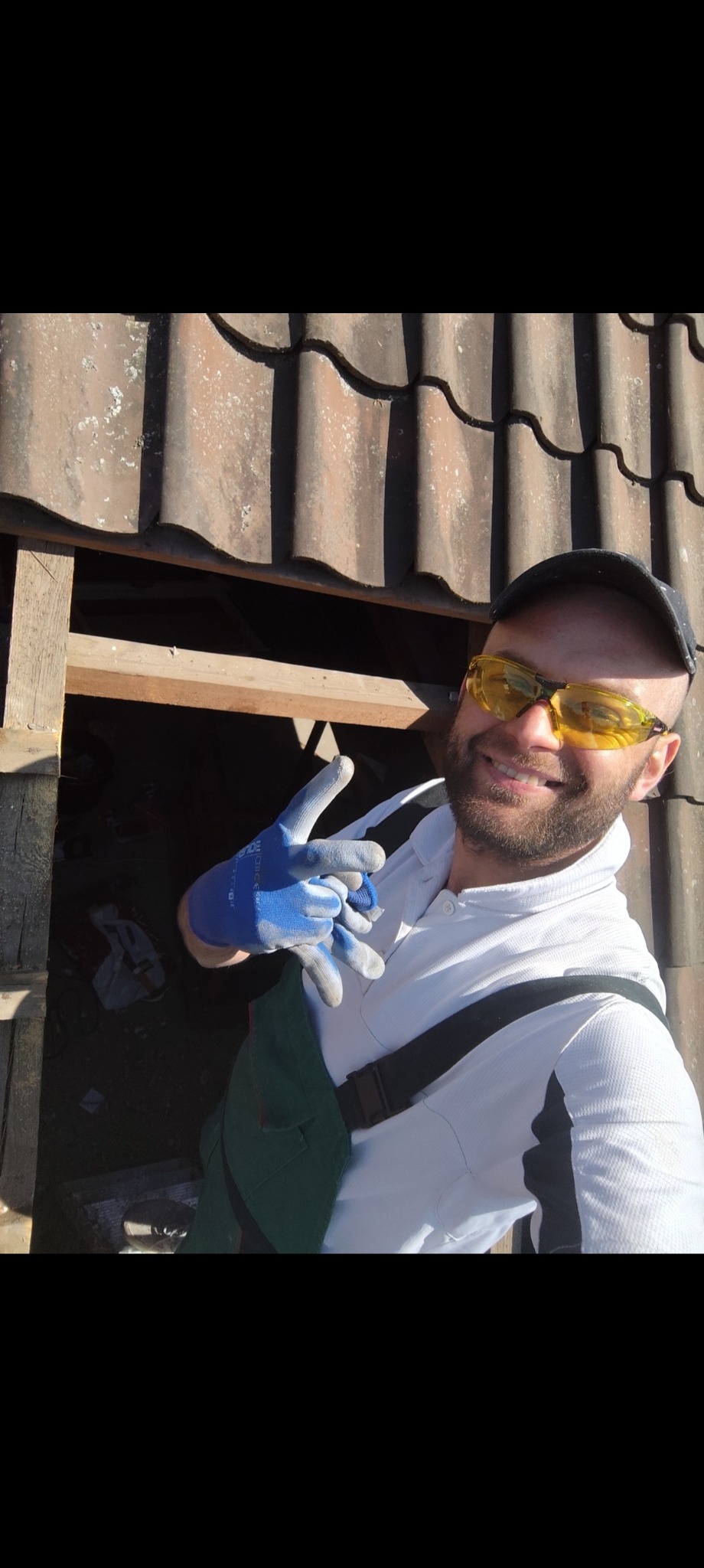 Uśmiechnięty dekarz w okularach ochronnych i roboczych rękawicach, na tle fragmentu dachu z brązowej dachówki podczas prac dekarskich.