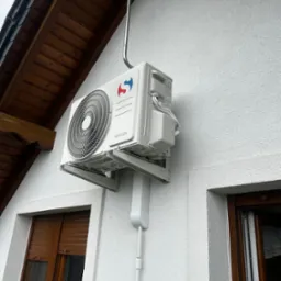 Biała jednostka zewnętrzna klimatyzacji Sinclair zamontowana na elewacji domu z drewnianym dachem i oknami z roletami zewnętrznymi. Widoczny przew&oacute;d odprowadzający.