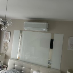 Peli-Cool klimatyzacja Dawid Bukowski - Biała klimatyzacja Samsung zamontowana nad łóżkiem w sypialni. Widoczny fragment lampy sufitowej i dekoracje ścienne. Minimalistyczny design wnętrza.