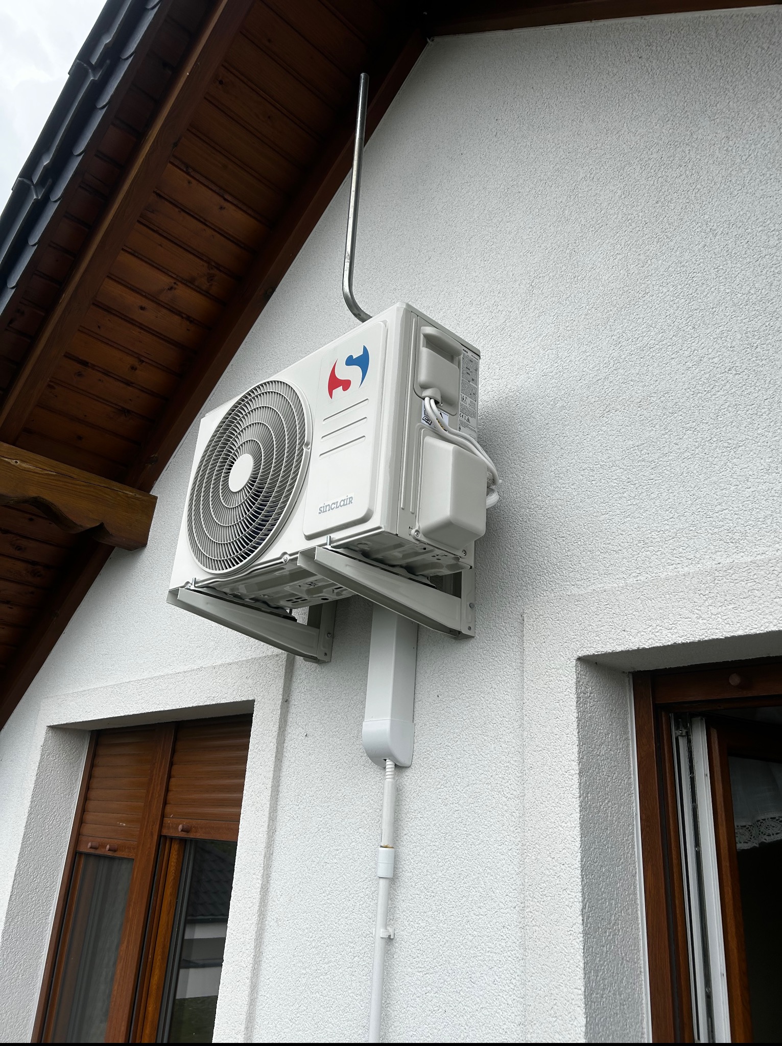 Biała jednostka zewnętrzna klimatyzacji Sinclair zamontowana na elewacji domu z drewnianym dachem i oknami z roletami zewnętrznymi. Widoczny przewód odprowadzający.