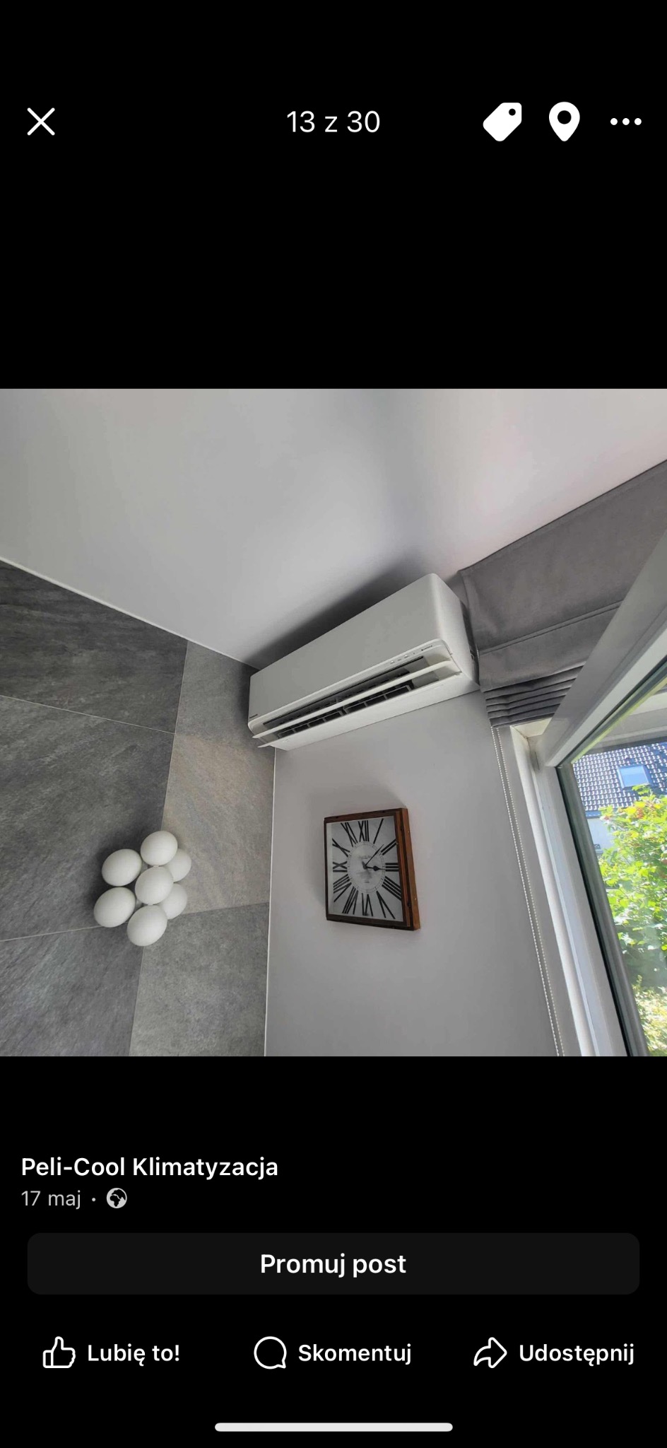 Biała klimatyzacja ścienna w nowoczesnym wnętrzu z szarą mozaiką, zegarem i dekoracją z białych kul. Widok z dołu, okno z roletą w tle.