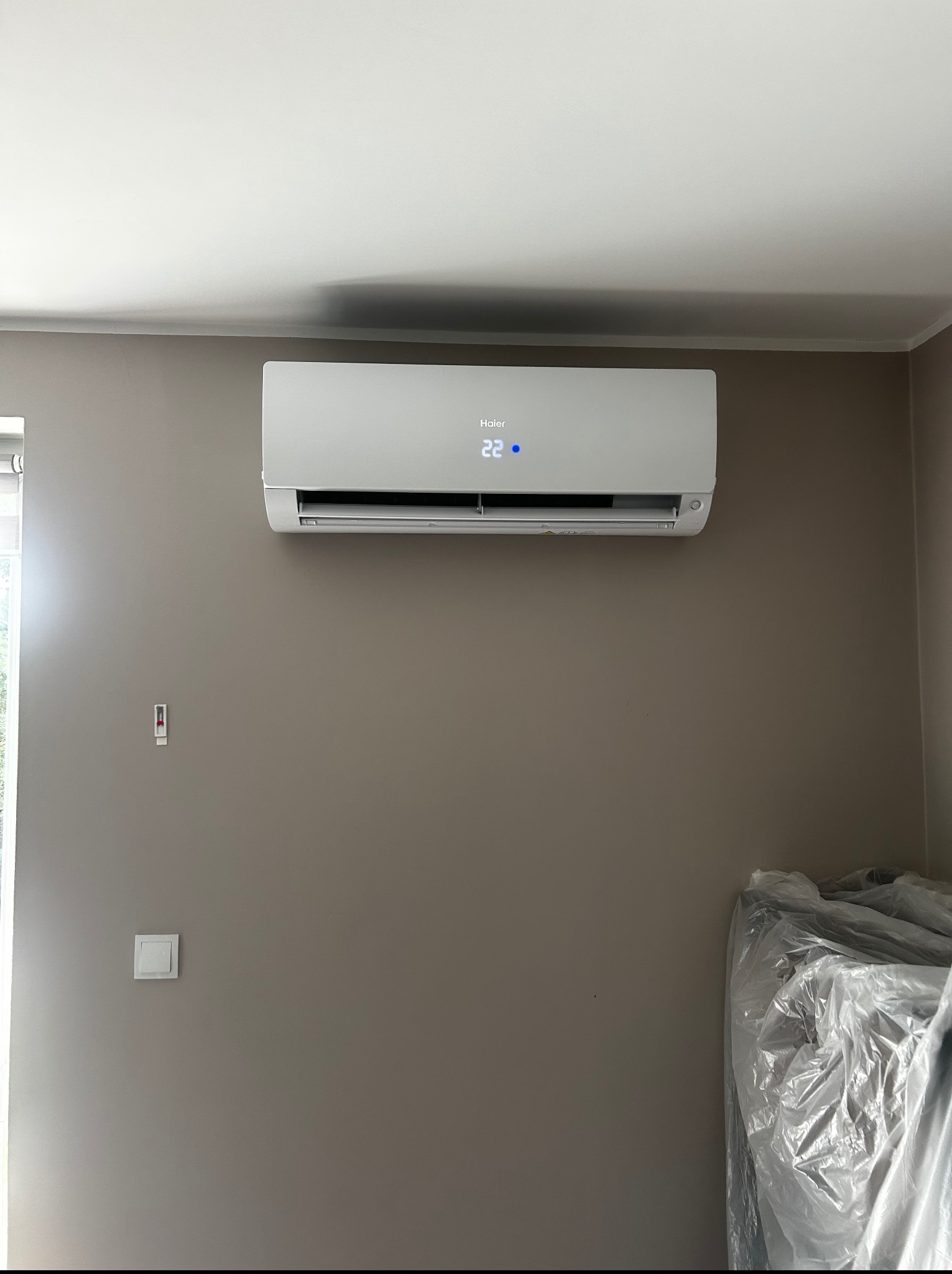 Zamontowany klimatyzator Haier na szarej ścianie w pomieszczeniu. Widoczny wyświetlacz z temperaturą 22 stopnie Celsjusza. Obok okno i zabezpieczone folią meble.