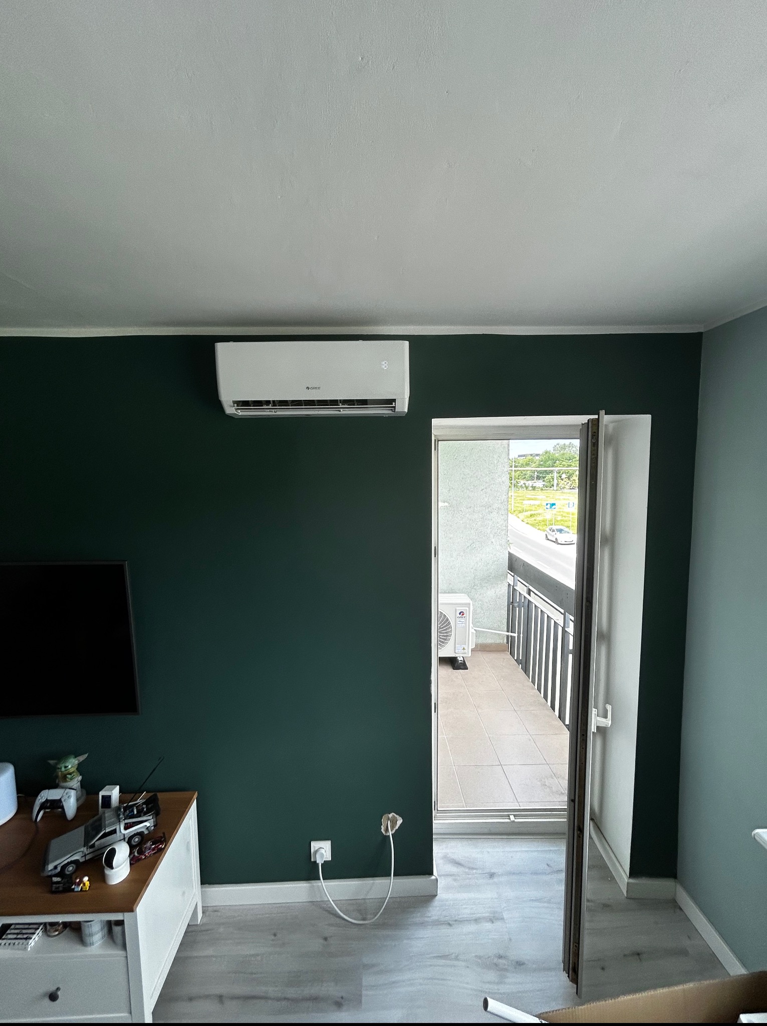 Nowoczesny klimatyzator Gree zamontowany na ciemnozielonej ścianie w salonie z wyjściem na balkon. Widoczny fragment telewizora i zabawki na komodzie.
