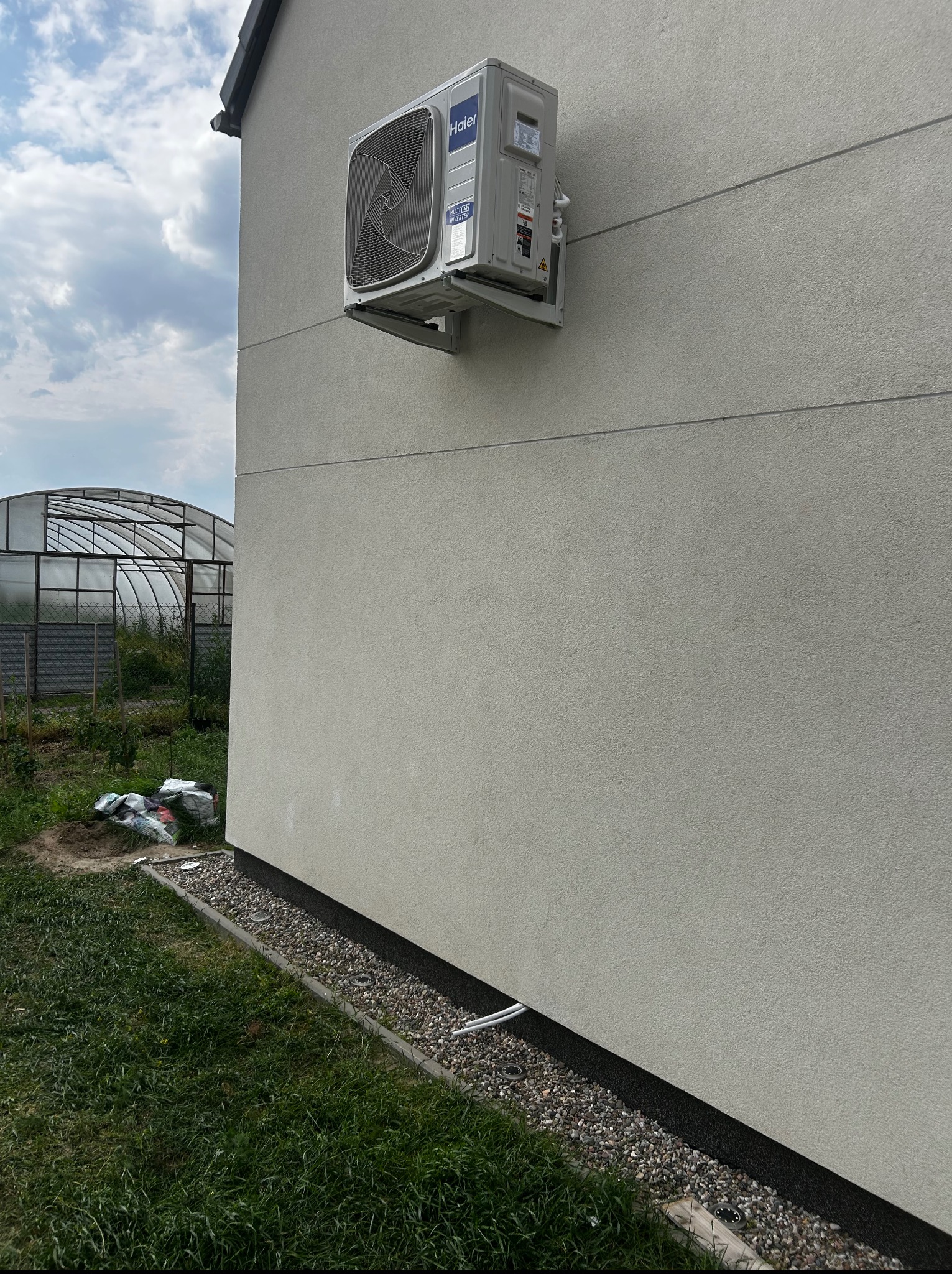 Zewnętrzna jednostka klimatyzacji Haier zamontowana na elewacji domu. Widoczne przewody i wsporniki. W tle szklarnia i trawnik. Ujęcie z dołu, podkreślające montaż.