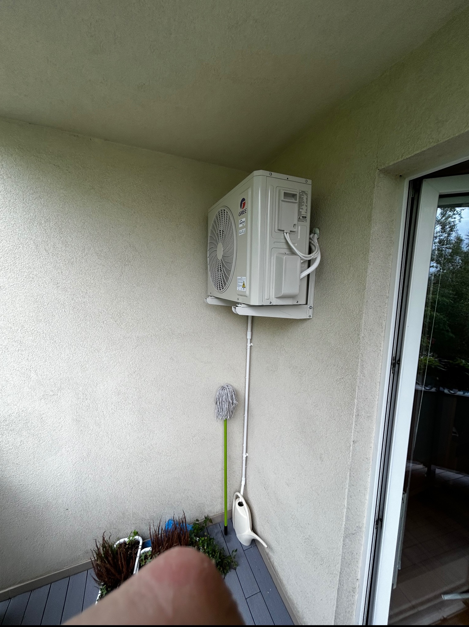 Klimatyzator marki Gree zamontowany na balkonie, z widoczną instalacją odprowadzającą skropliny. Obok rośliny w doniczce i mop. Widok z dołu na urządzenie.
