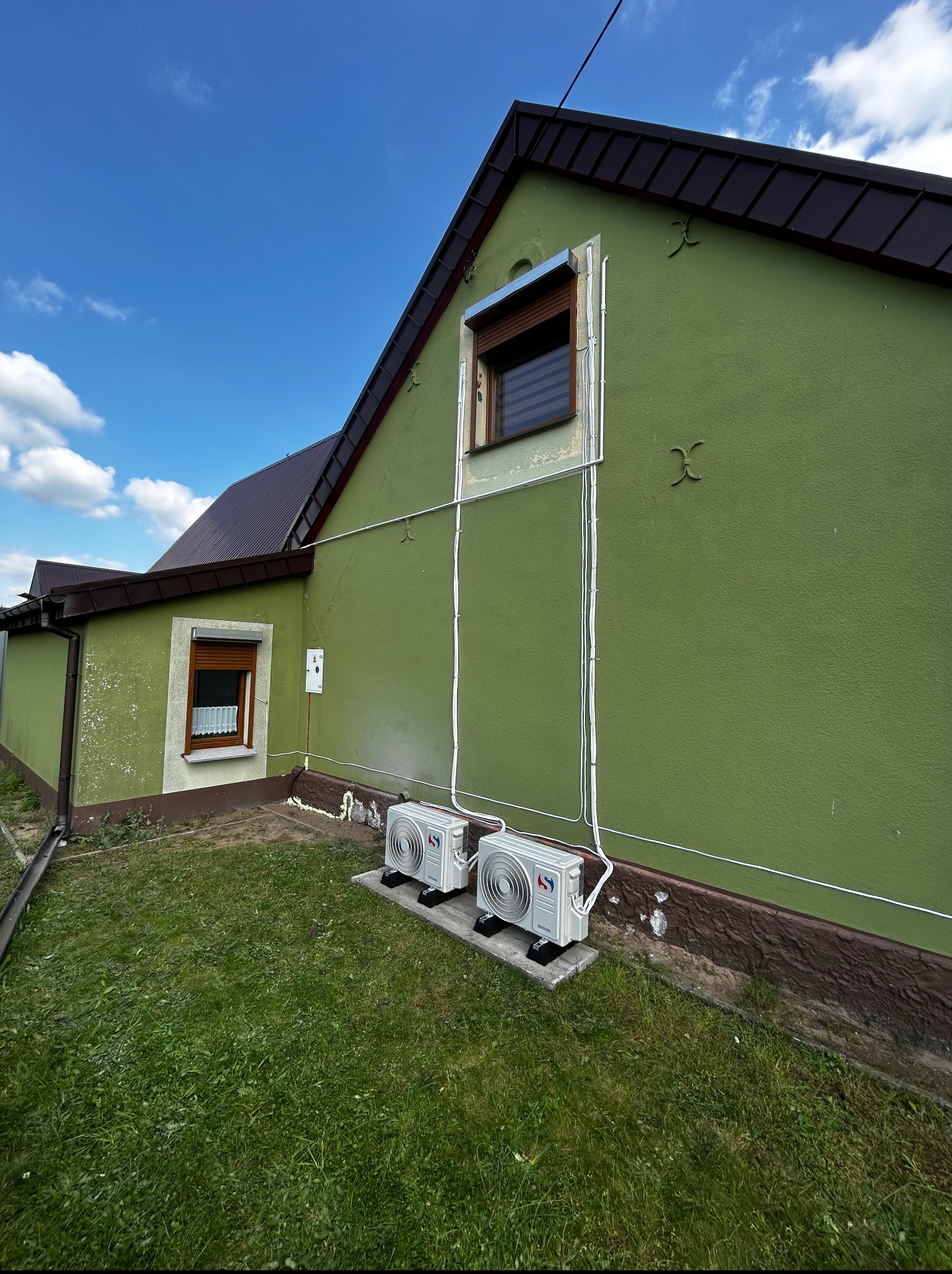 Dwa klimatyzatory na zewnątrz zielonego domu z brązowym dachem, widoczne przewody instalacji i część trawnika. Błękitne niebo w tle.