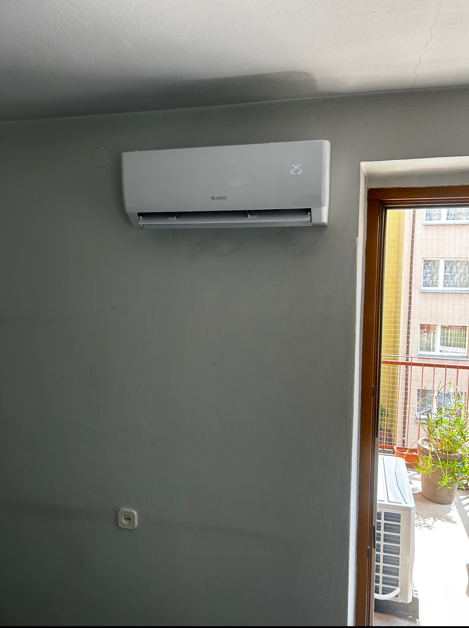 Biały klimatyzator Gree zamontowany na szarej ścianie, z wyświetlaczem temperatury 25 stopni. Widok na balkon przez otwarte drzwi.