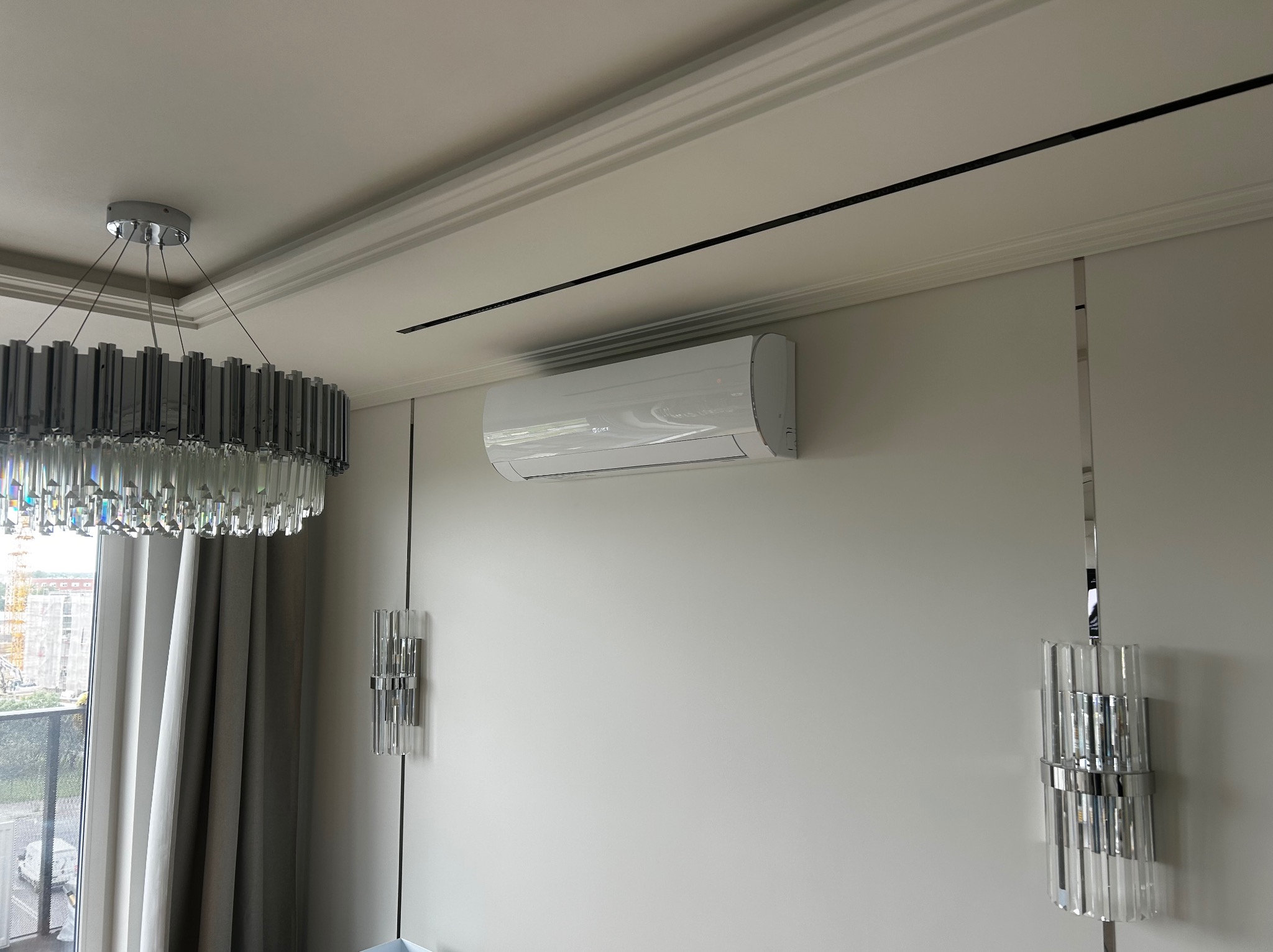 Biała klimatyzacja typu split zamontowana na ścianie w nowoczesnym wnętrzu z designerskim oświetleniem i dekoracyjnymi listwami sufitowymi.